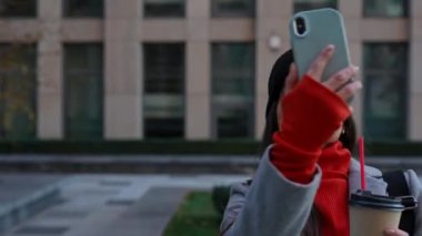 Şık bir genç kadın akıllı telefondan video görüşmesi yapıyor. Sıcak palto ve şapka giyen kadın, bir sonbahar günü şehir manzarası arasında yürürken video aracılığıyla iletişim kuruyor. Metropolis 'te sonbahar günü.