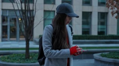 Akıllı telefondan mesaj atan ya da göz gezdiren şık bir genç kadın. Paltosu ve şapkası olan bir kadın telefonda megapolis 'in çok katlı bir şehir binasının arka planında iletişim kuruyor..