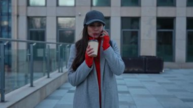 Akıllı telefondan konuşan şık bir genç kadın. Sıcak bir palto ve şapka giymiş bir kadın telefonda çok katlı bir şehir binasının arka planında sohbet ediyor..