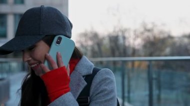 Akıllı telefondan konuşan şık bir genç kadın. Sıcak bir palto ve şapka giymiş bir kadın telefonda çok katlı bir şehir binasının arka planında sohbet ediyor..