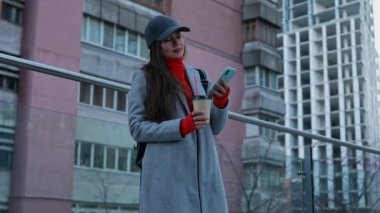 Akıllı telefondan mesaj atan ya da göz gezdiren şık bir genç kadın. Paltosu ve şapkası olan bir kadın telefonda megapolis 'in çok katlı bir şehir binasının arka planında iletişim kuruyor..