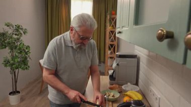 Son sınıf öğrencisinin hayatında bir gün. Yaşlı gri saçlı adam sabah mutfakta, sağlıklı kahvaltı hazırlıyor, muz kesiyor ve yulaf ezmesiyle kaseye ekliyor, pozitif ifade. Günlük rutin yaşam tarzı kavramı