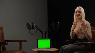 Çevrimiçi podcast gösterisi. Kadın konuk podcast stüdyosunda şık deri koltukta oturuyor, röportajı kaydediyor, dijital içerik yaratıyor, kameraya konuşuyor ve dizüstü bilgisayarında kroma tuşlu yeşil ekran kullanıyor..