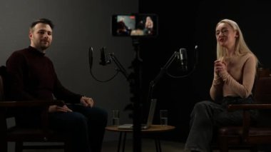 Çevrimiçi podcast gösterisi. Erkek sunucu ve kadın konuk sanatçı podcast stüdyosunda mikrofon ve akıllı telefon kullanarak dijital içerik yaratarak, konuşarak ve seyirciyi selamlayarak röportaj yapıyorlar..