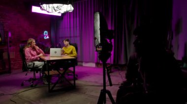 Çevrimiçi yayın. Çatı katındaki podcast stüdyosundaki röportajın sahne arkası, dijital içerik yaratma, sunucu ve misafir konuşması, adam krom anahtar yeşil ekran maketli akıllı telefonu gösteriyor, kameraman yan tarafta oturuyor.