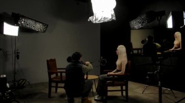 Çevrimiçi podcast gösterisi. Sahne arkası çekimlerinde kadın konuk şık podcast stüdyosunda röportaj yapmaya hazırlanıyor ses mühendisi mikrofonları tamir ediyor, yan tarafta video akışı var..