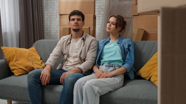 Yeni eve taşınma konsepti. Genç çift yeni daireye taşınıyor, yer karton kutularla dolu, erkek ve kadın yeni dairenin odasındaki kanepede oturuyor, olumlu bir ifade tarzı düşünüyorlar..