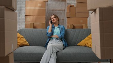 Yeni eve taşınma konsepti. Yeni daireye taşınan genç bir çift, karton kutularla dolu bir yer, akıllı telefondaki yeni apartman sohbetlerinin olduğu koltukta oturan bir kadın, mutlu bir pozitif ifade..