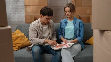 Yeni eve taşınma konsepti. Genç çift yeni daireye taşınıyor, yer karton kutularla dolu, erkek ve kadın kanepede oturuyor, renk paletinde oda yenileme renklerini seçiyorlar..