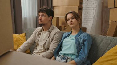 Yeni eve taşınma konsepti. Genç çift yeni daireye taşınıyor, yer karton kutularla dolu, erkek ve kadın yeni dairenin odasındaki kanepede oturuyor, olumlu bir ifade tarzı düşünüyorlar..