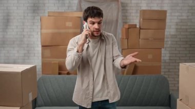 Yeni eve taşınma konsepti. Genç çift yeni daireye taşınıyor, yer karton kutularla dolu, adam yeni dairenin odasında duruyor ve akıllı telefondan konuşuyor, hayal kırıklığına uğramış olumsuz ifade..