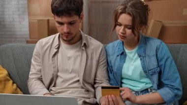 Yeni eve taşınma konsepti. Genç çift yeni bir daireye taşınıyor, yer karton kutularla dolu, erkek ve kadın kanepede oturuyor dizüstü bilgisayardaki kart numarası, internetten heyecanlı bir ifade satın alıyorlar..