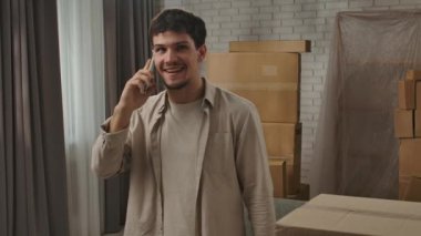 Yeni eve taşınma konsepti. Genç çift yeni daireye taşınıyor, yer karton kutularla dolu, adam yeni dairenin odasında duruyor ve akıllı telefondan konuşuyor, mutlu pozitif ifade..