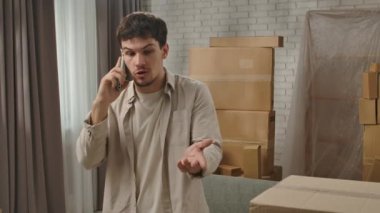 Yeni eve taşınma konsepti. Genç çift yeni daireye taşınıyor, yer karton kutularla dolu, adam yeni dairenin odasında duruyor ve akıllı telefondan konuşuyor, hayal kırıklığına uğramış olumsuz ifade..