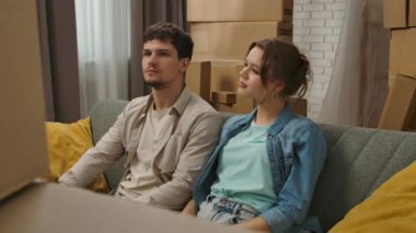 Yeni eve taşınma konsepti. Genç çift yeni daireye taşınıyor, yer karton kutularla dolu, erkek ve kadın yeni dairenin kanepesinde oturuyor, etrafa olumlu bir ifadeyle bakınıyorlar..