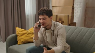 Yeni eve taşınma konsepti. Yeni daireye taşınan genç bir çift, karton kutularla dolu bir yer, akıllı telefondaki yeni apartman sohbetlerinin olduğu koltukta oturan adam, mutlu bir pozitif ifade..