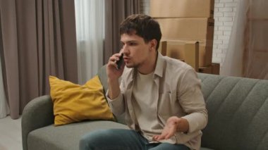 Yeni eve taşınma konsepti. Yeni daireye taşınan genç bir çift, karton kutularla dolu bir yer, yeni dairedeki kanepede oturan bir adam ve akıllı telefondan konuşan, hayal kırıklığına uğramış negatif ifade..