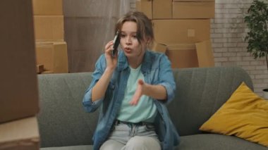 Yeni eve taşınma konsepti. Yeni daireye taşınan genç bir çift, karton kutularla dolu bir yer, yeni dairedeki kanepede oturan bir kadın ve akıllı telefondan konuşan, hayal kırıklığına uğramış negatif ifade.