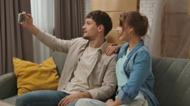 Yeni eve taşınma konsepti. Yeni daireye taşınan genç bir çift, karton kutularla dolu bir yer, kanepede oturan erkek ve kadın akıllı telefondan video konuşmaları, mutlu bir pozitif ifade..
