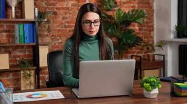 Ofiste çalışan bir iş adamı. Genç iş kadını geç saatlere kadar ofis dairesinde çalışıyor, dizüstü bilgisayar kullanıyor, klavyede yazmakla meşgul, kameraya bakıyor ve gülümsüyor, olumlu ifade veriyor..