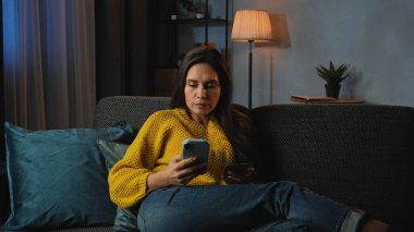 Günlük rutin yaşam tarzı konsepti. Akşamları işten sonra evde dinlenen, oturma odasında kanepede oturup televizyon izleyen, akıllı telefon uygulamasında kart numarası yazan, internetten alışveriş yapan genç bir kadın..