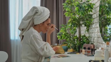 Günlük rutin yaşam tarzı konsepti. Sabahları genç esmer kadın aynanın karşısına oturur yatak odasında bornozla ve kafasında havluyla, günlük makyajını yapar, alnına fırçayla gölgeler sürer..