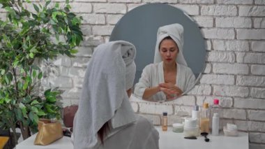 Günlük rutin yaşam tarzı konsepti. Sabahları genç bir kadın aynanın karşısına oturur yatak odasında bornozlu ve kafasında havluyla, ellerine krem sürerek, kaşlarına da fırça sürerek. Arka plan..