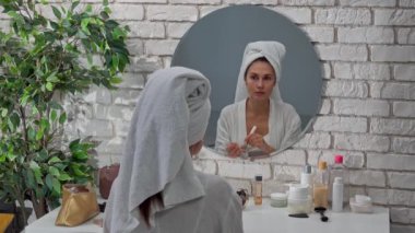 Günlük rutin yaşam tarzı konsepti. Sabahları genç esmer kadın aynanın önünde yatak odasında bornozlu ve kafasında havluyla oturuyor, yüzüne büyük bir fırça sürerek makyaj pudrası uyguluyor. Arka plan..