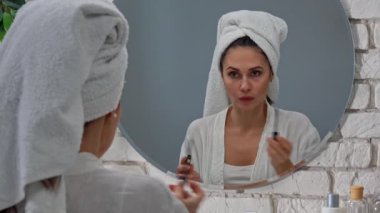 Günlük rutin yaşam tarzı konsepti. Sabahları genç bir kadın aynanın karşısına oturur yatak odasında bornozlu ve kafasında havluyla kirpikleri için maskara ve kaşlarına kozmetik ürünler sürerek. Arka plan..