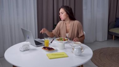 Günlük rutin yaşam tarzı konsepti. Sabahları genç esmer kadın, oturma odasında sağlıklı bir kahvaltı yapıyor, masada oturuyor, salata yiyor ve dizüstü bilgisayarını kontrol ediyor..