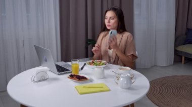 Günlük rutin yaşam tarzı konsepti. Sabahları genç esmer kadın, oturma odasında sağlıklı bir kahvaltı yapıyor, masada oturuyor, yemek yiyor ve bilgisayarla çalışıyor, akıllı telefondan konuşuyor..