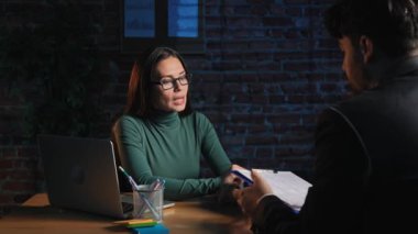 Günlük rutin yaşam tarzı konsepti. Ofisteki genç esmer kadın gece geç saatlere kadar çalışıyor, iş kadını masasında erkek müşteriyle konuşuyor, kontrat kağıtlarını okuyor ve imzalıyor..
