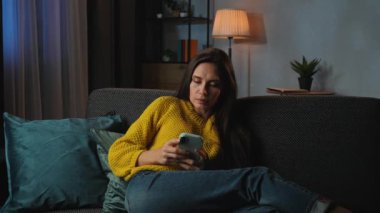 Günlük rutin yaşam tarzı konsepti. Akşamları işten sonra evde kanepede dinlenen, televizyon izleyen, akıllı telefondan mesaj atan, iyi haberler alan, gülümseyen ve dans eden genç bir kadın..
