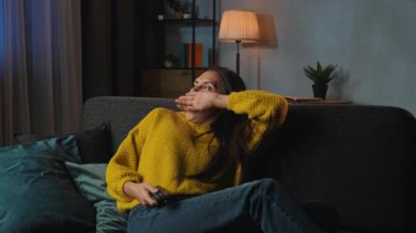 Günlük rutin yaşam tarzı konsepti. Akşam işten sonra genç bir kadın evde dinleniyor, kanepede oturuyor televizyon seyrediyor, kanalları değiştiriyor yoruluyor ve sıkılıyor, uykulu bir yüz ifadesi takınıyor.