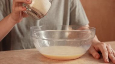 Günlük ev yemeği rutini. Kadın evde, mutfakta masada oturuyor ve waffle kahvaltısı için hamur hazırlıyor, karıştırılmış yumurtalarla kaseye bir bardak şeker ekliyor. Yavaş çekim.