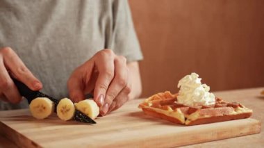 Günlük ev yemeği rutini. Kadın evde, mutfakta masada oturuyor ve waffle kahvaltısı için hamur hazırlıyor, fırında waffle için tahtaya bıçakla muz kesiyor. Yavaş çekim.