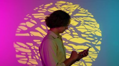 Renkli stüdyo geçmişinde telefonu olan biri. Gündelik kıyafetlerle gezen bir adamın portresi akıllı telefondan kayıyor, spot ışıkları altında dikiliyor, pembe mavi neon üzerinde izole edilmiş, stüdyo arkaplanı var..