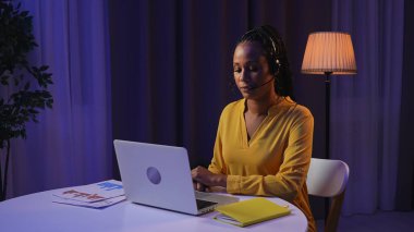 Günlük rutin yaşam tarzı konsepti. Akşamları geç saatlere kadar çalışan, dizüstü bilgisayar kullanan, kulaklık takan, konuşan, klavyede yazan genç Afrikalı Amerikalı kadın.,