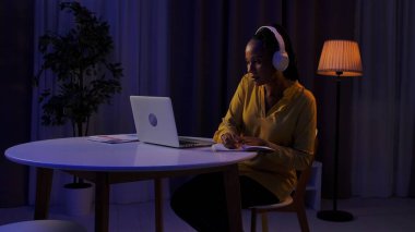 Günlük rutin yaşam tarzı konsepti. Akşamları geç saatlere kadar çalışan, kulaklık takan, dizüstü bilgisayarda müzik dinleyen ve deftere yazı yazan Afro-Amerikan genç bir kadın..