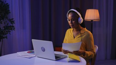Günlük rutin yaşam tarzı konsepti. Akşamları geç saatlere kadar çalışan, kulaklık takmış, bilgisayardan video konuşmaları yapan genç Afrikalı Amerikalı bir kadın..