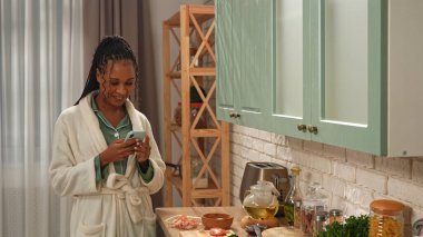 Günlük rutin yaşam tarzı konsepti. Sabahları mutfakta sağlıklı kahvaltı hazırlayan, akıllı telefondan mesaj atan, mesajlaşan genç Afrikalı Amerikalı kadın..