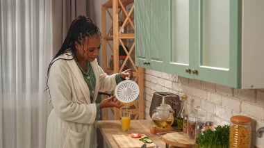 Günlük rutin yaşam tarzı konsepti. Genç Afro-Amerikalı kadın sabah mutfakta sağlıklı bir kahvaltı hazırlıyor, kadın bardağa taze portakal suyu dolduruyor..