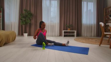 Günlük rutin yaşam tarzı konsepti. Genç Afrikalı Amerikalı kadın sabahları yatak odasında ısınıyor yoga minderine oturuyor ve yerde egzersiz yapmaya başlıyor, bacaklarını ve bacaklarını esnetiyor..