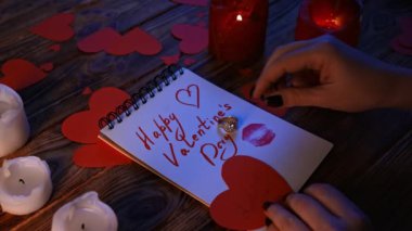 Sevgililer Günü konsepti. Ahşap masa, yanan mumlar ve dağınık kağıt kalpler, mutlu sevgililer günü yazılı not defteri masaya uzanmış, kadın parmağına elmas yüzük takıyor..