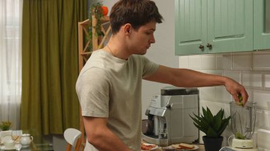 Günlük rutin yaşam tarzı konsepti. Genç adam sabah mutfakta sağlıklı bir kahvaltı hazırlıyor, taze meyve suyu hazırlıyor, blender 'a kivi dilimleri atıyor..