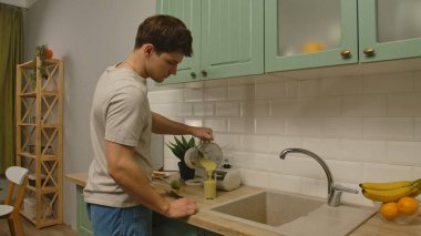 Günlük rutin yaşam tarzı konsepti. Sabahleyin genç adam mutfakta sağlıklı bir kahvaltı hazırlıyor, taze meyve suyu hazırlıyor, bardaktan su dolduruyor..
