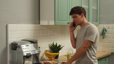 Günlük rutin yaşam tarzı konsepti. Sabahları mutfakta sağlıklı bir kahvaltı hazırlayan, yemek hazırlayan ve akıllı telefondan konuşan genç bir adam..