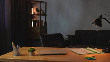 Kapalı dizüstü bilgisayarı, belgeleri ve kırtasiyesi olan ahşap bir masası olan şık bir ofis. Siyah bir masa lambası odaklanmış ışık sağlarken, dekoratif bir raf sıcaklık ve karakter ekler..