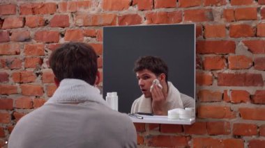 Günlük rutin yaşam tarzı konsepti. Sabah genç bir adam aynanın karşısında duruyor banyoda boynunda havluyla, günlük hijyen rutini yapıyor, yüzüne tıraş köpüğü sürüyor..