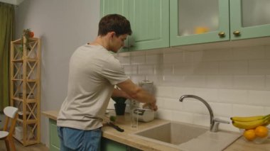 Günlük rutin yaşam tarzı konsepti. Genç adam sabah mutfakta sağlıklı bir kahvaltı hazırlıyor, taze smoothie hazırlıyor, karıştırıcıdan bardağa su dolduruyor ve bir yudum alıyor..